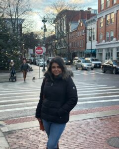 A woman in a black winter coat stands on Essex Street in Salem, Massachusetts, with holiday lights on a tree, brick buildings, and cars in the background. Pedestrians and a dog are crossing the street.