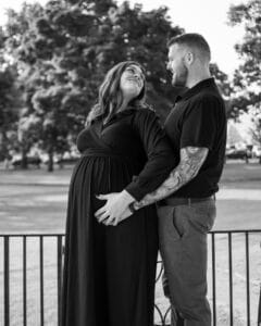 A black-and-white photo of a pregnant woman in a flowing black dress and her partner embracing from behind, both smiling lovingly at each other in a peaceful Salem park.