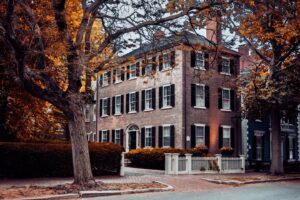 A historic brick colonial house with black shutters and a white fence, framed by colorful fall leaves in Salem, Massachusetts.