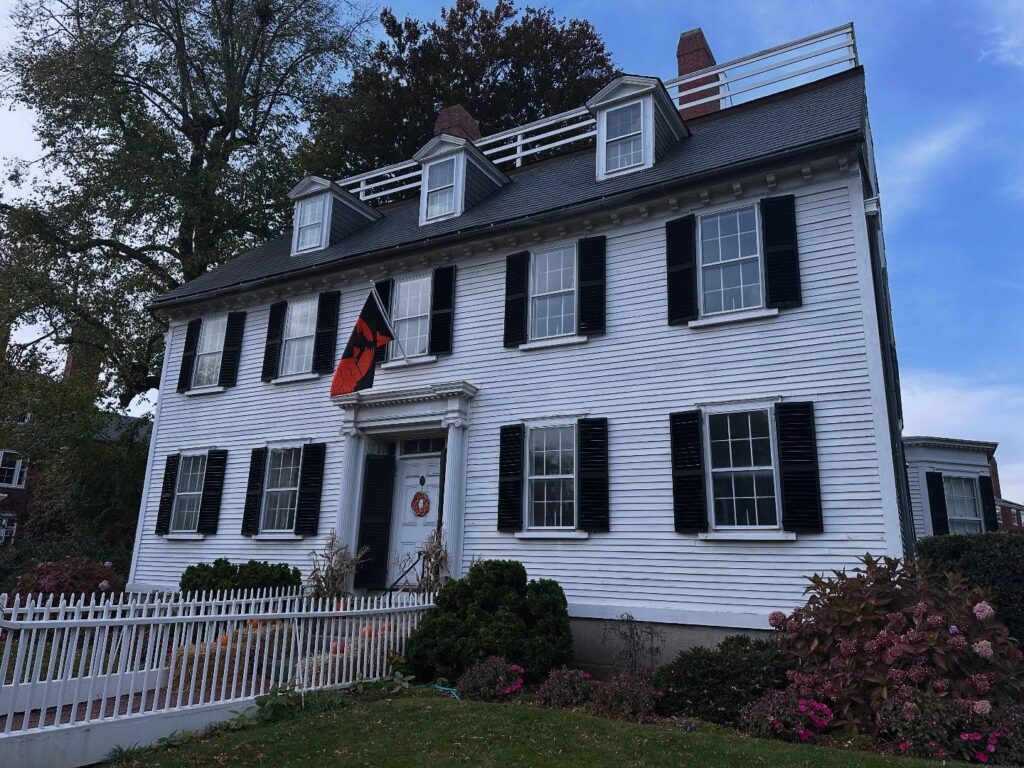 The historic Ropes Mansion in Salem, Massachusetts, a white colonial-style house with black shutters, surrounded by vibrant fall foliage and a white picket fence.