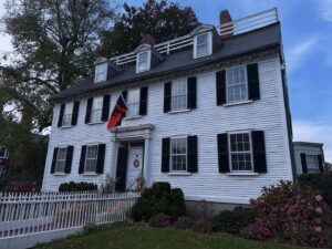 The historic Ropes Mansion in Salem, Massachusetts, a white colonial-style house with black shutters, surrounded by vibrant fall foliage and a white picket fence.