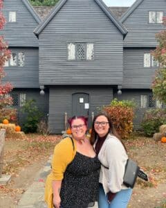Two friends pose happily in front of the Witch House in Salem, Massachusetts, surrounded by vibrant autumn foliage, pumpkins, and hay bales that enhance the seasonal charm of this historic landmark.