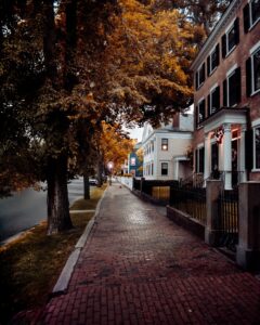 A quiet brick-paved sidewalk in Salem, Massachusetts, lined with historic homes and tall trees glowing with vibrant autumn leaves.