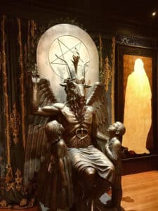 The iconic Baphomet statue at the Salem Satanic Temple in Massachusetts, featuring intricate details and two children gazing up at the seated figure against a pentagram backdrop.