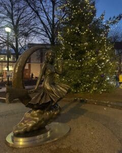 The Bewitched statue in Salem, Massachusetts, shows a figure sitting on a broomstick in front of a large Christmas tree decorated with twinkling lights and ornaments during twilight.