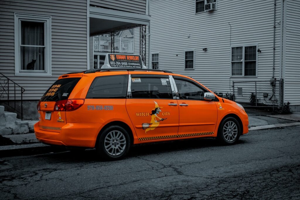 An orange Witch City taxi with a witch logo on its side is parked in Salem, Massachusetts, while a black cat peers out from a nearby window.
