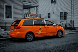 An orange Witch City taxi with a witch logo on its side is parked in Salem, Massachusetts, while a black cat peers out from a nearby window.