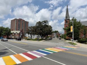 A rainbow crosswalk in Salem, Massachusetts, connecting to a peaceful street with historic buildings and a tall church steeple under a sunny sky.