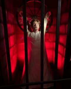 A lifelike figure of a person bound to a wheel, enclosed in a red-lit cage, showcased at the Salem Museum of Torture to portray historical punishment methods.