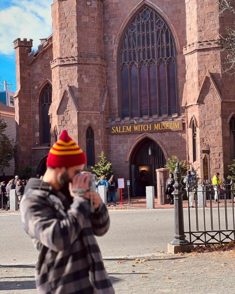 A man in a red and yellow beanie takes a photo in front of the Salem Witch Museum in Salem, Massachusetts. The gothic stone building with arched windows and the museum sign stands in the background with visitors gathered near the entrance.