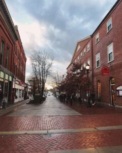 A view of Central Street in Salem, Massachusetts, lined with brick buildings and lampposts. The wet pavement reflects the soft glow of the evening light.
