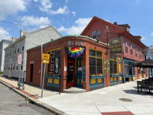Mercy Tavern in Salem, Massachusetts, with its vibrant red brick exterior, colorful accents, and a pride flag hanging above the entrance, on a sunny day.