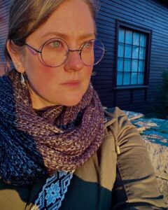 A close-up of a woman in Salem, Massachusetts, wearing glasses, a scarf, and a jacket, illuminated by warm golden hour sunlight with a historic wooden building in the background.
