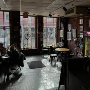 Inside Front Street Coffeehouse in Salem, Massachusetts, featuring large windows with heart decorations, warm brick walls, and local artwork. A few people are enjoying drinks and conversation at wooden tables. Natural light streams in, creating a relaxed and inviting atmosphere.