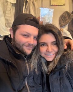 A couple dressed in warm winter clothing smiles for a selfie indoors in Salem, Massachusetts. The background features rustic decor with historical fabrics and artifacts.