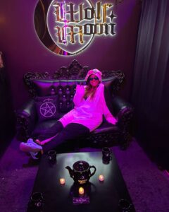 A person in a playful onesie relaxes on a gothic black couch at Coven in Salem, Massachusetts, under a glowing "Wolf Moon" sign, surrounded by witchy decor and purple lighting.
