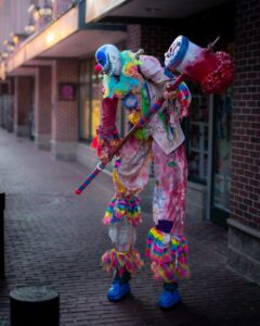 A scary clown in colorful, tattered clothes holding a giant mallet walks along the brick streets of Salem, Massachusetts, adding a spooky and theatrical vibe to the scene.
