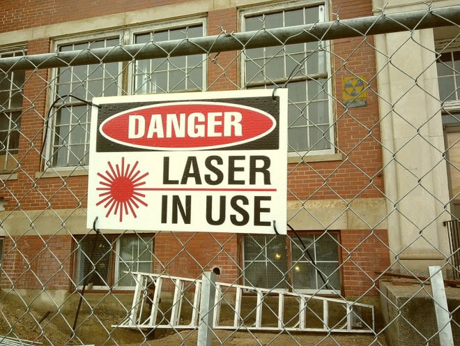 A warning sign reading "Danger: Laser in Use" attached to a chain-link fence in front of the historic St. Marys School building in Salem, Massachusetts.