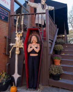 A woman poses inside a red coffin labeled "The Haunted Neighborhood" outside a spooky-themed display in Salem, Massachusetts. Skeletons, a pumpkin, and other Halloween decorations complete the eerie yet playful setting.