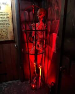 A skeleton in a tall iron cage, illuminated with ominous red lighting, is displayed at the Salem Museum of Torture to depict historical methods of confinement.