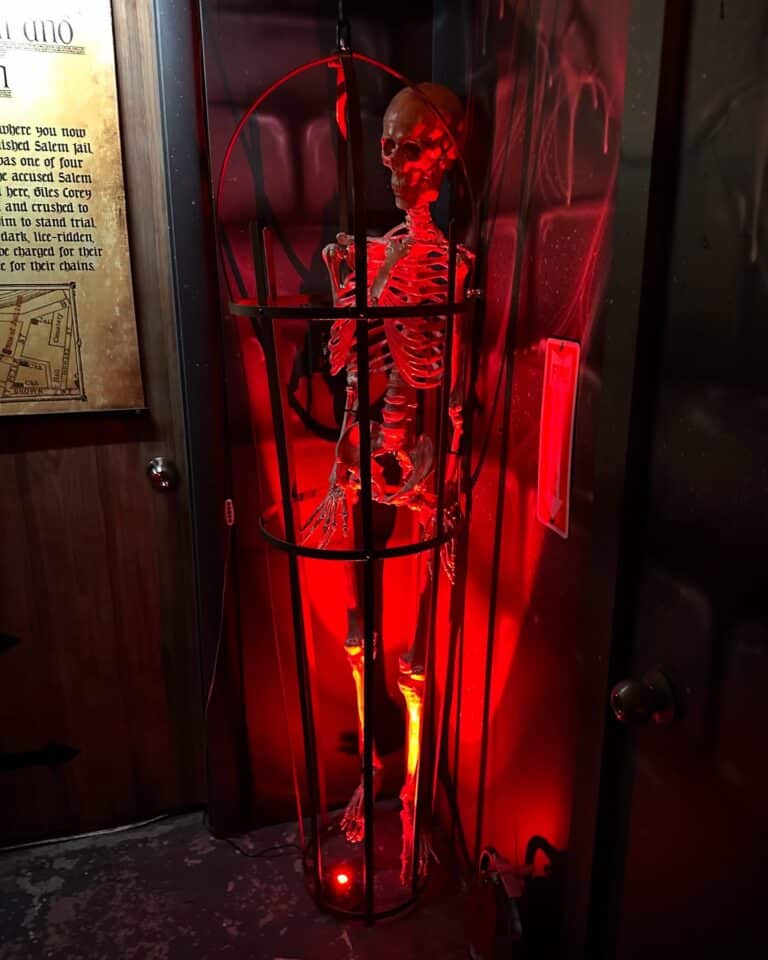 A skeleton in a tall iron cage, illuminated with ominous red lighting, is displayed at the Salem Museum of Torture to depict historical methods of confinement.