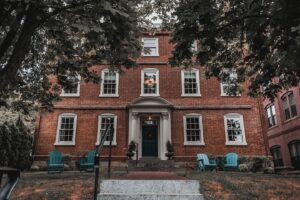 A picturesque view of The Merchant Hotel in Salem, Massachusetts, showcasing its historic red brick exterior surrounded by trees and a cozy seating area with Adirondack chairs on the front lawn.