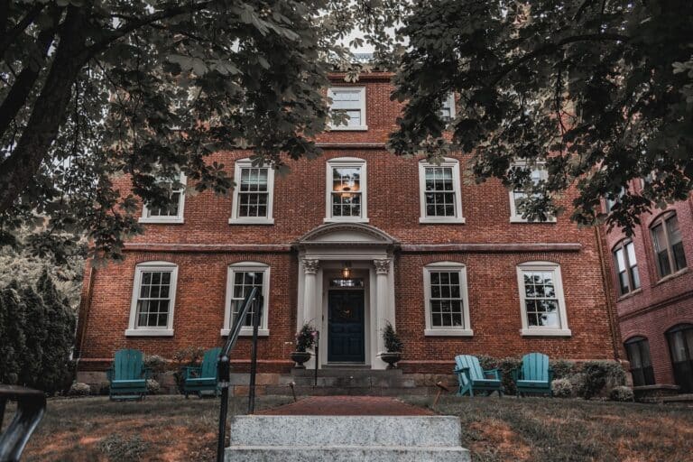 A picturesque view of The Merchant Hotel in Salem, Massachusetts, showcasing its historic red brick exterior surrounded by trees and a cozy seating area with Adirondack chairs on the front lawn.