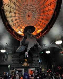 A suspended witch sculpture flying on a broomstick beneath an orange-lit dome at Blackcraft in Salem, Massachusetts, surrounded by gothic architecture.