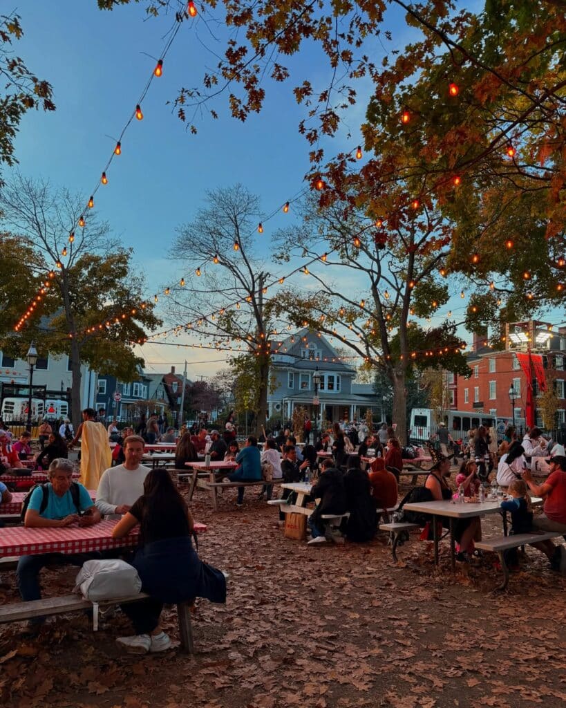 A lively gathering at Salem Common in Salem Massachusetts during fall, with people sitting at picnic tables under strings of glowing lights and autumn leaves covering the ground.