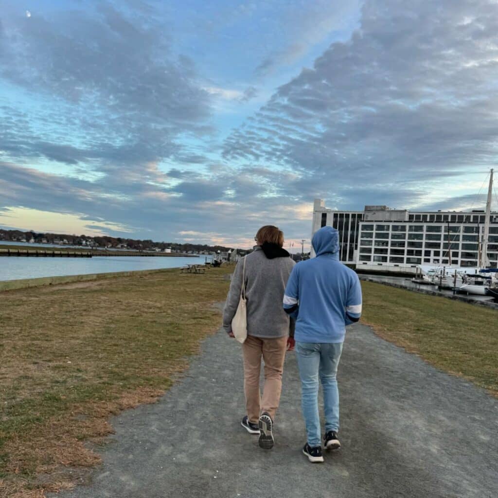 Two people walking along a gravel path by the water in Salem, Massachusetts during sunset. A marina and modern building are visible to the right, with sailboats docked nearby.