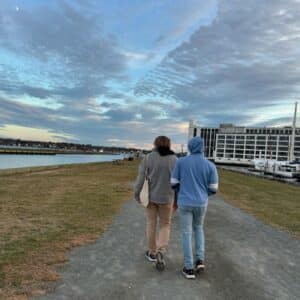 Two people walking along a gravel path by the water in Salem, Massachusetts during sunset. A marina and modern building are visible to the right, with sailboats docked nearby.