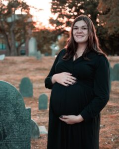 A pregnant woman in a black dress stands smiling in a historic graveyard at sunset, surrounded by gravestones with warm, golden light filtering through the trees.