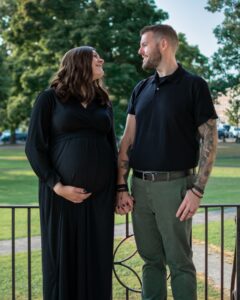 A glowing pregnant woman in a long black dress and her partner in a black polo and green pants hold hands and smile at each other in a serene park in Salem, Massachusetts.