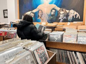 A person browsing through vinyl records in a Salem, Massachusetts record store, surrounded by shelves of albums and a vibrant mural featuring famous composers in the background.