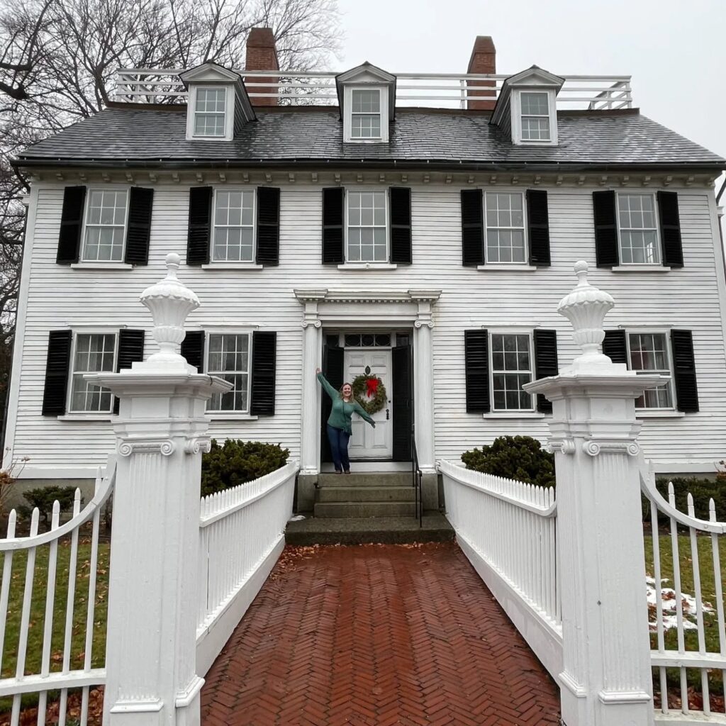 A person stands joyfully at the entrance of the Ropes Mansion in Salem, Massachusetts, decorated with a holiday wreath on the door and surrounded by white fencing and a brick walkway.