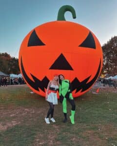 Two friends in creative costumes pose in front of a giant inflatable jack-o'-lantern at Salem Common during a Halloween celebration.