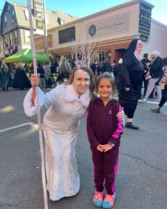 A woman dressed as an icy queen with a staff poses with a smiling young girl on Essex Street in Salem, Massachusetts, during a lively Halloween celebration.