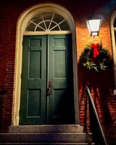 A green wooden door with a semicircular window at the top, located at the Old Town Hall in Salem, Massachusetts, adorned with a holiday wreath featuring a red bow and illuminated by a warm lantern.