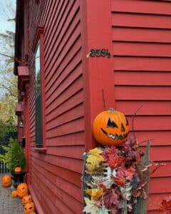 A vibrant red building in Salem, Massachusetts with the address "358½" displayed on the corner. A cheerful jack-o'-lantern sits atop a skeleton figure draped with colorful autumn leaves, surrounded by more pumpkins in a festive Halloween setup.