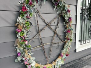 A large wreath featuring a pentagram design made from twigs, adorned with greenery and vibrant pink and purple flowers, hanging on a dark gray wall.