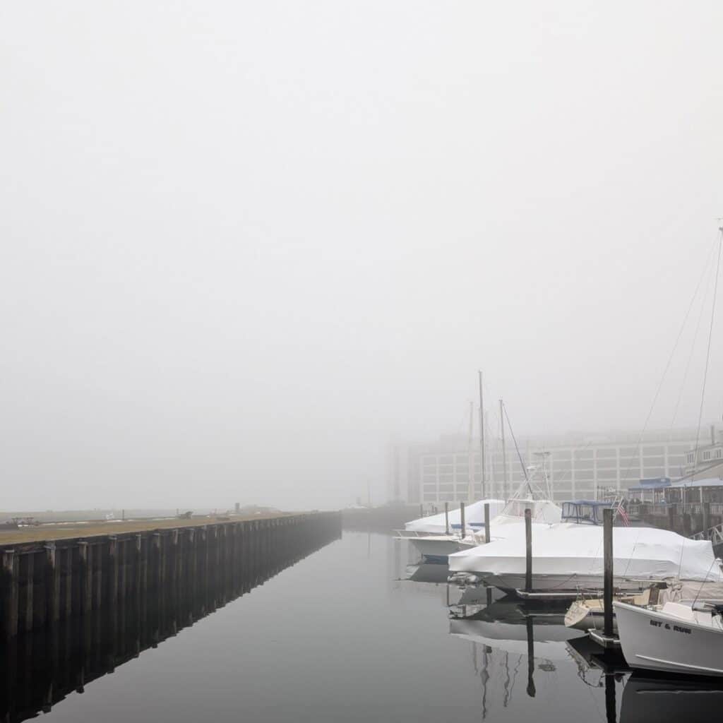 Boats docked along the calm waters of the Salem waterfront on a foggy morning, with the mist obscuring buildings and creating a serene atmosphere.