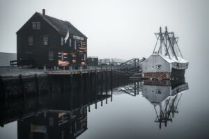 A serene foggy scene at Salem Maritime National Historic Site in Salem, Massachusetts, featuring the historic Sail Loft building and a docked replica ship partially covered with tarps, reflecting in the calm water.