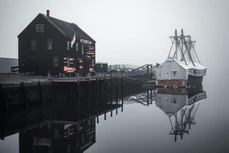 The Friendship of Salem ship docked beside the historic black Sail Loft building in Salem, Massachusetts, reflected in calm, foggy waters.