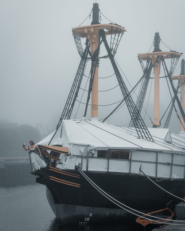 A misty morning view of The Friendship of Salem docked at Derby Wharf in Salem, Massachusetts, with its tall masts and intricate rigging standing out against the foggy sky.