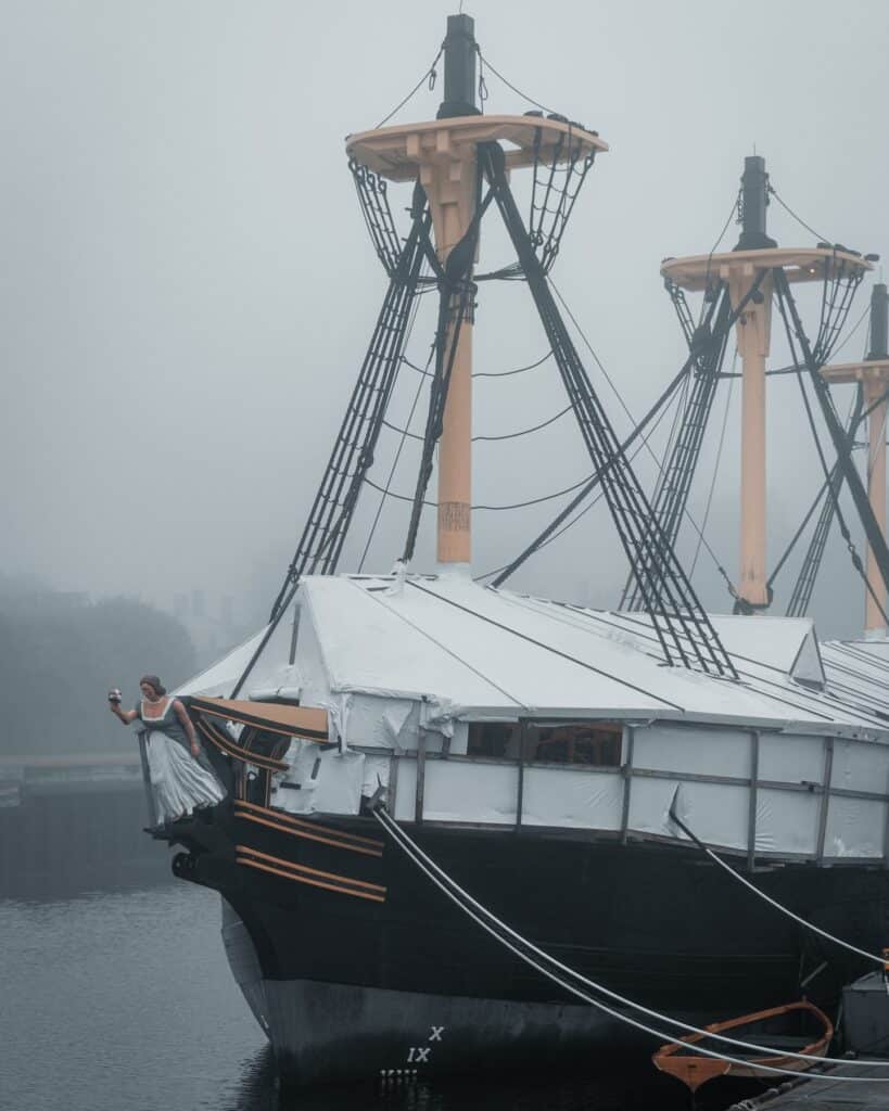 A misty morning view of The Friendship of Salem docked at Derby Wharf in Salem, Massachusetts, with its tall masts and intricate rigging standing out against the foggy sky.