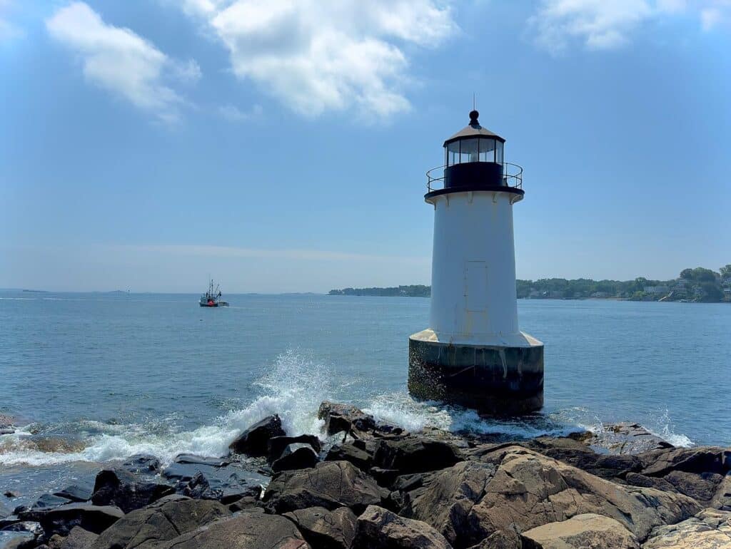 Fort Pickering Lighthouse in Salem, Massachusetts, standing on a rocky shore with ocean waves splashing and a small fishing boat in the distance under a sunny blue sky.