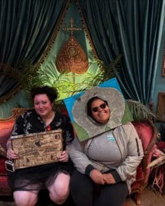 Two individuals seated inside the Salem Witch Board Museum in Salem, Massachusetts. One holds a vintage Ouija board, while the other wears a playful costume piece resembling a painted stone. They are surrounded by rich decor featuring velvet drapes and a decorative Ouija board sign.