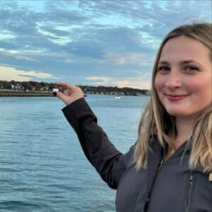 A woman near Derby Light Station in Salem, Massachusetts, smiles as she holds her hand up to make it look like she's pinching a distant boat on the water. The calm harbor, shoreline houses, and a soft sky create a peaceful background.