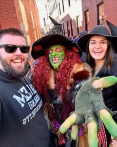 A cheerful group selfie with Borah the witch in Salem, Massachusetts, featuring her green face, red hair, and outstretched hand, surrounded by brick buildings and excited visitors.
