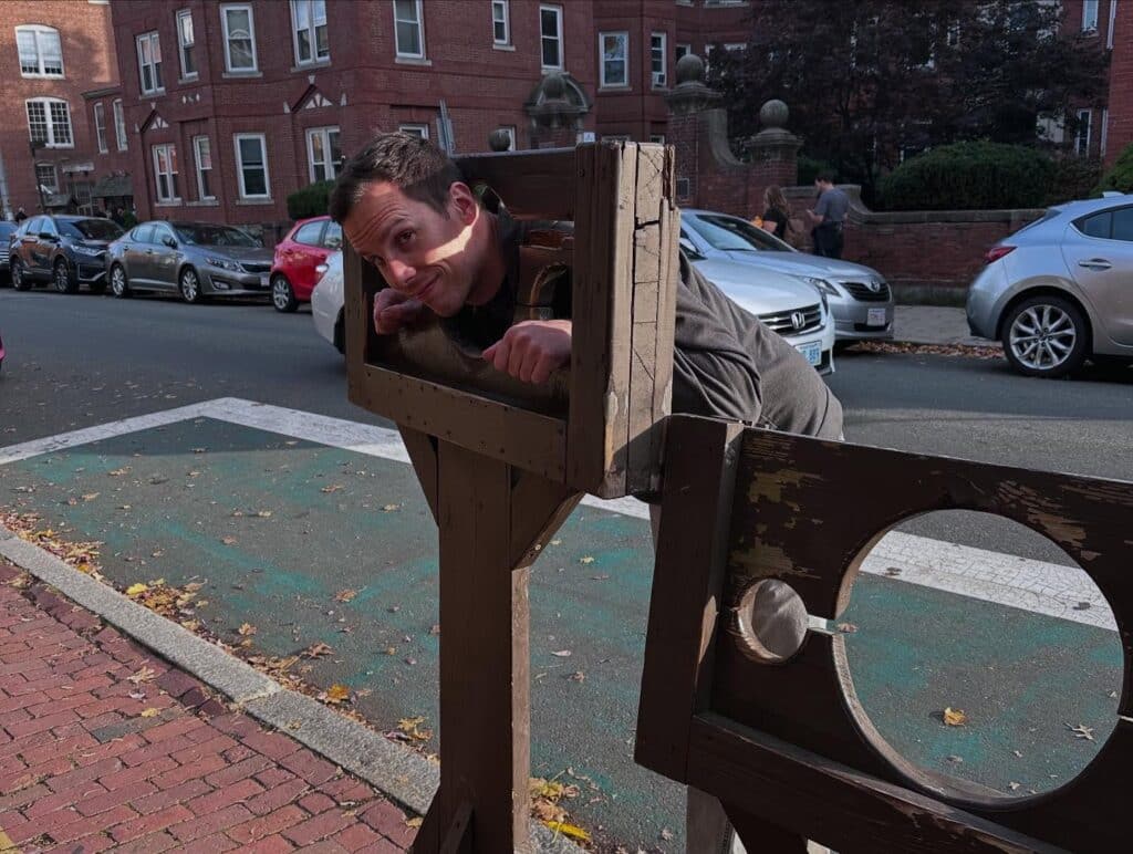 A playful moment at the Witch Dungeon Museum in Salem, Massachusetts, with a man leaning through a wooden stockade, surrounded by a backdrop of brick buildings and parked cars.